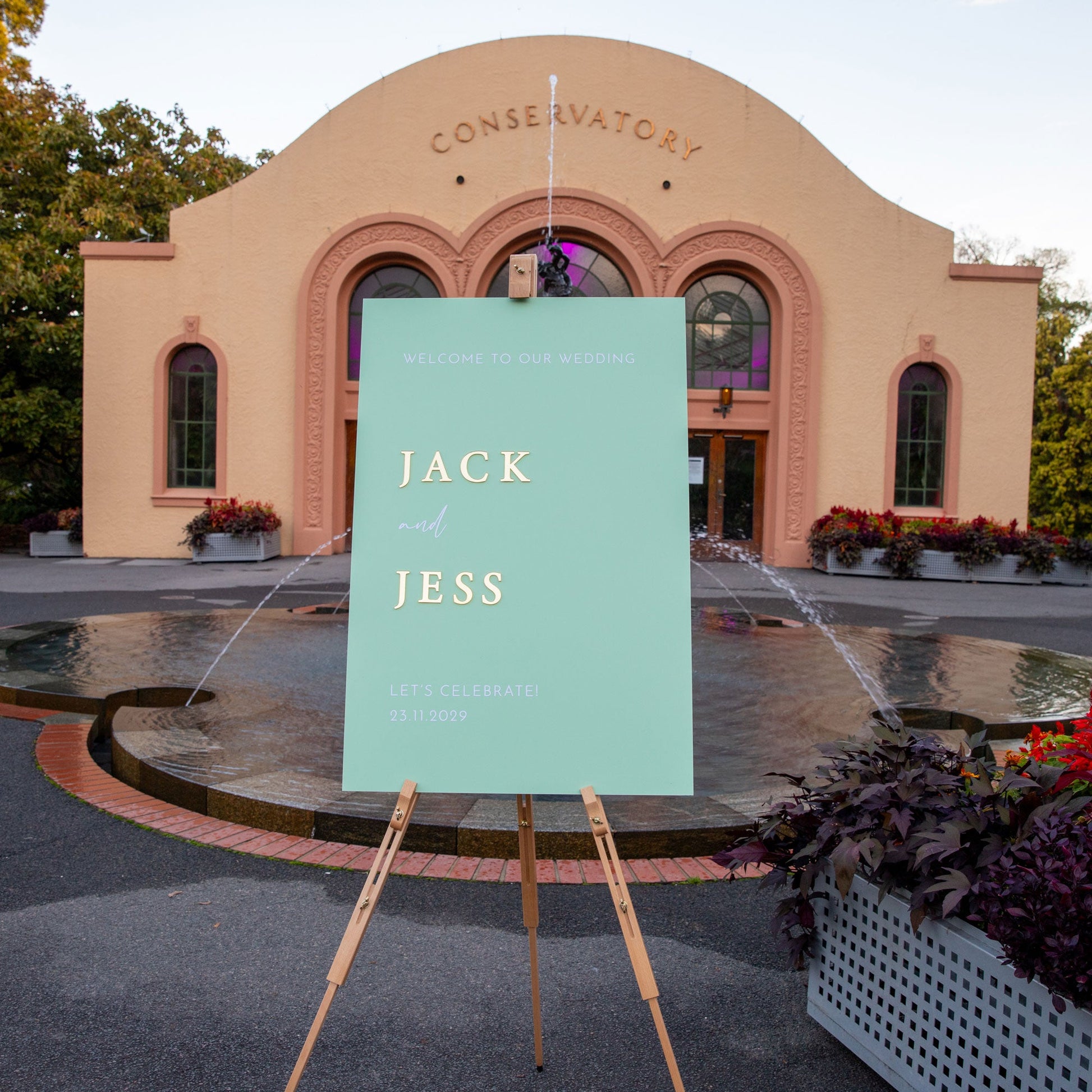 Personalised wedding sign featuring sage green acrylic and 3D gold names, set outside a venue with fountain.